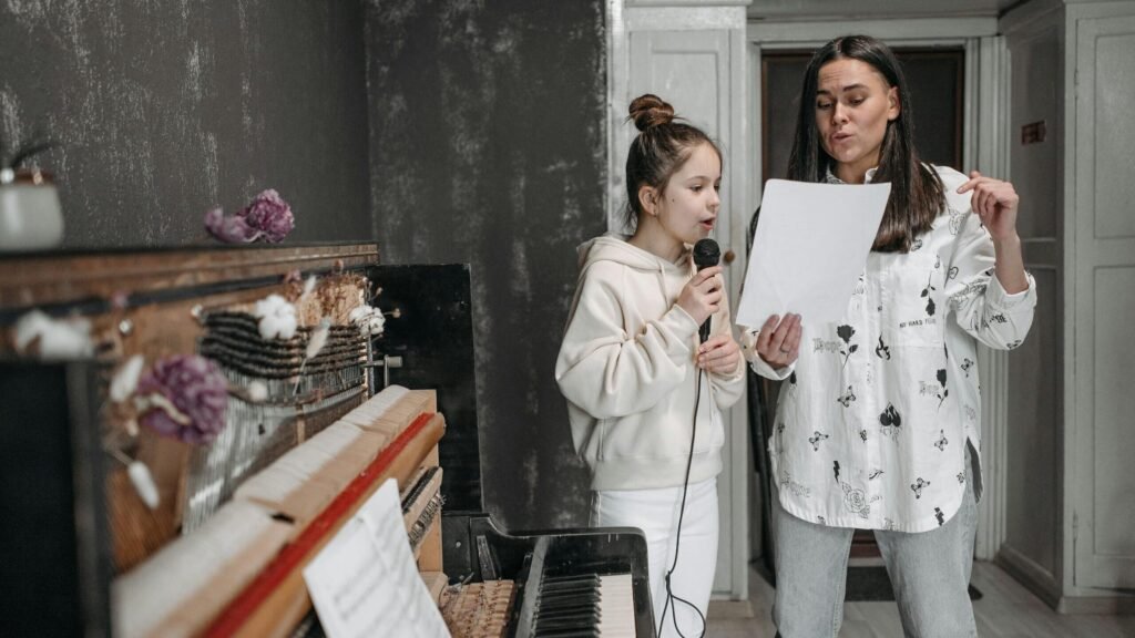 A girl taking a voice lesson from her teacher, singing into a microphone by a piano.